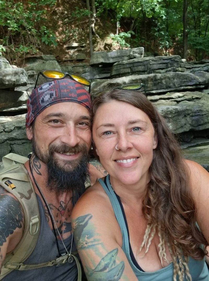 Brad and Jenny, the husband-and-wife owners of Natural State Cleanout Co., smiling together in front of a rocky creek bed in the woods