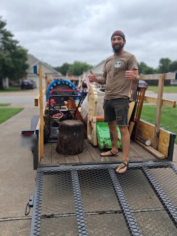 Brad giving two thumbs up from a trailer loaded with a lawn mower, a tree stump, a kids' playhouse and a wooden crib